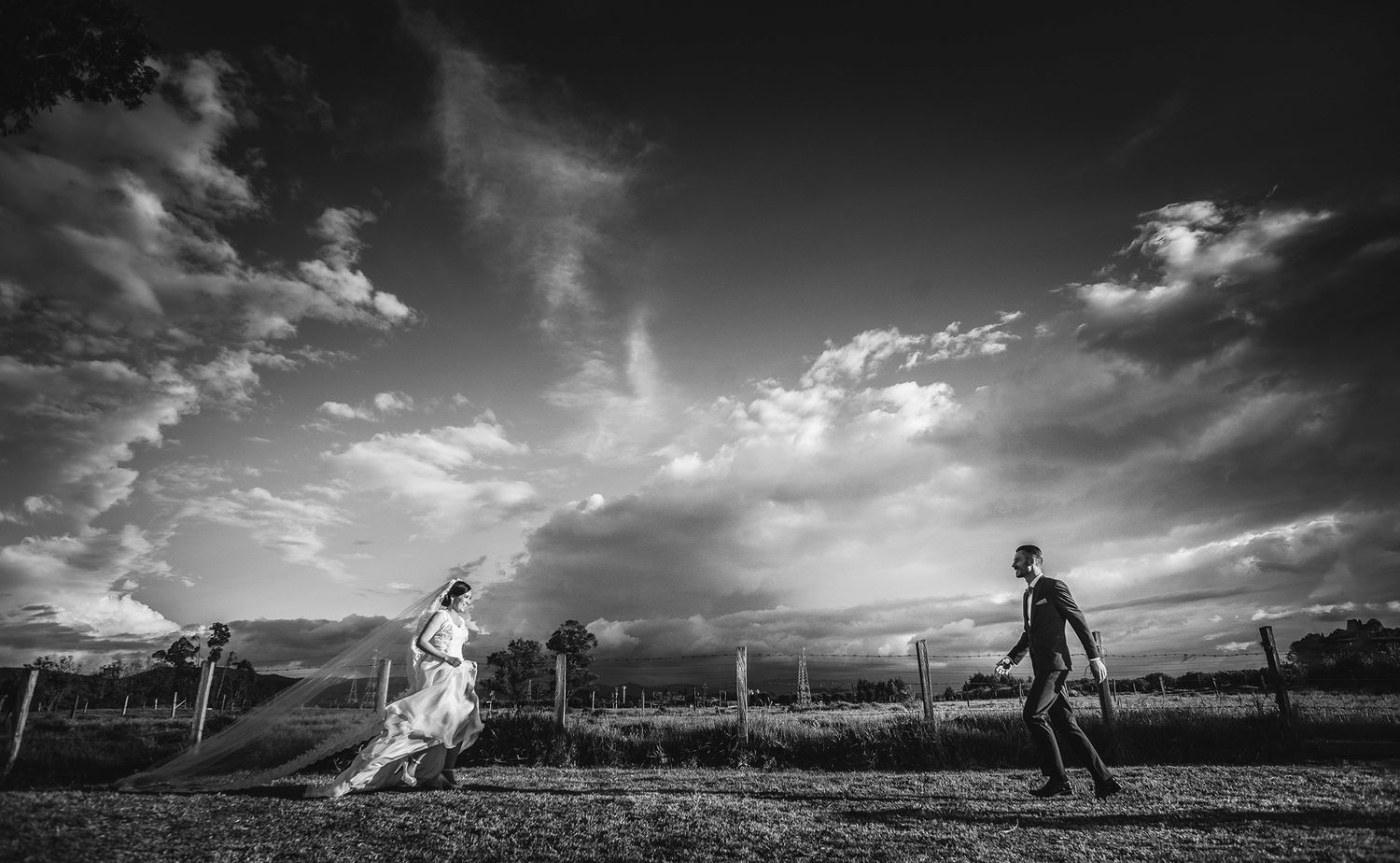 Black and white photo of a bride and groom walking through a field under dramatic cloudy skies.