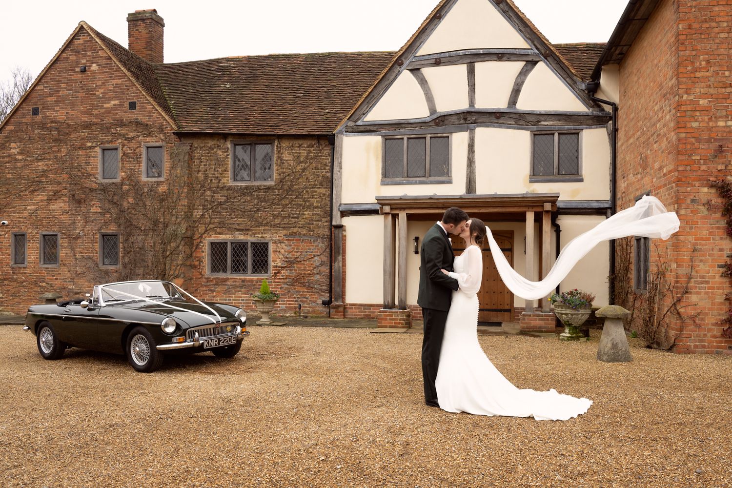 Bride and groom kissing outside a Tudor manor house with a classic black convertible car on their wedding day.