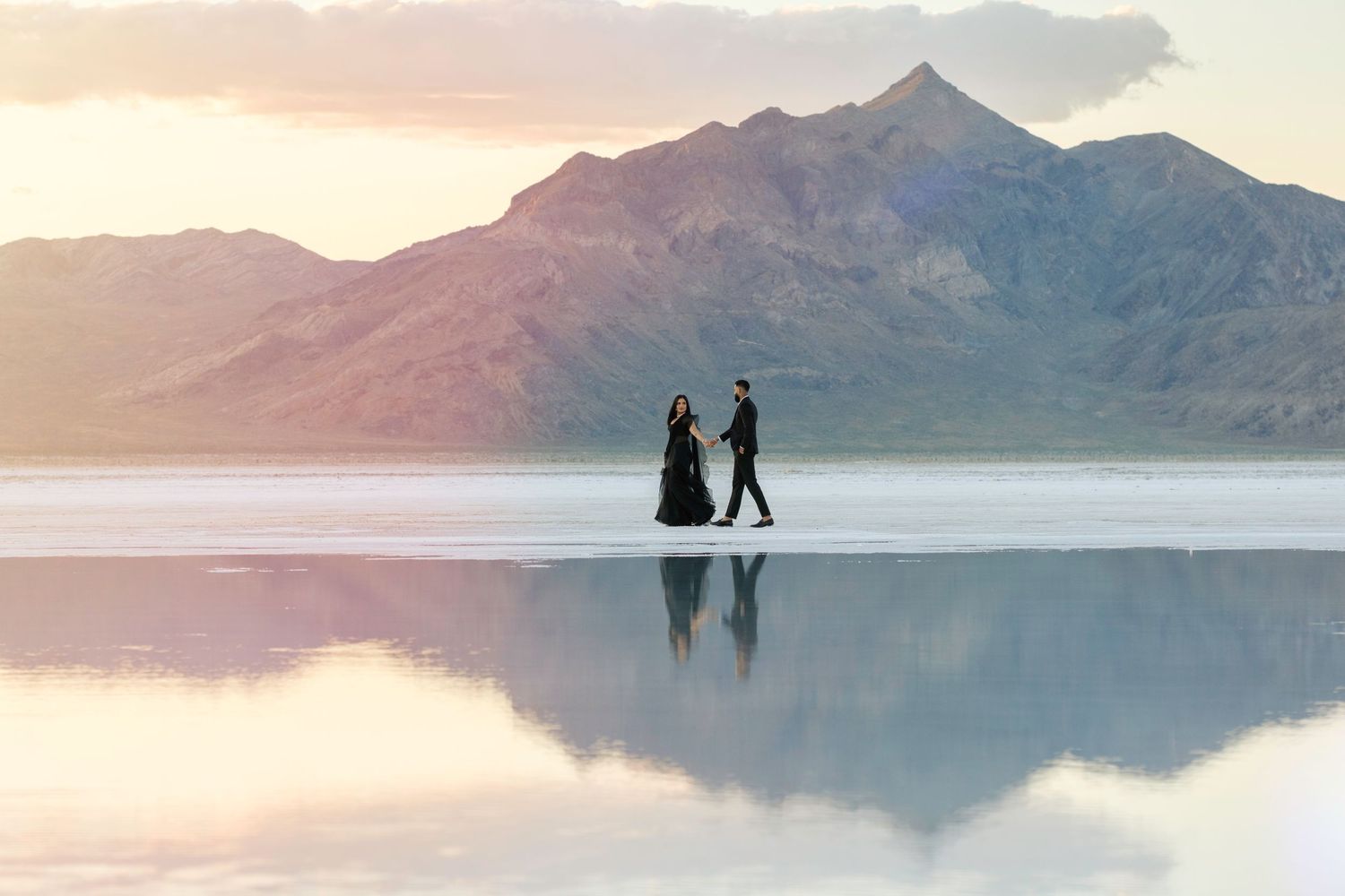 Couple in black attire walking on reflective Bonneville Salt Flats with mountain backdrop at sunset.