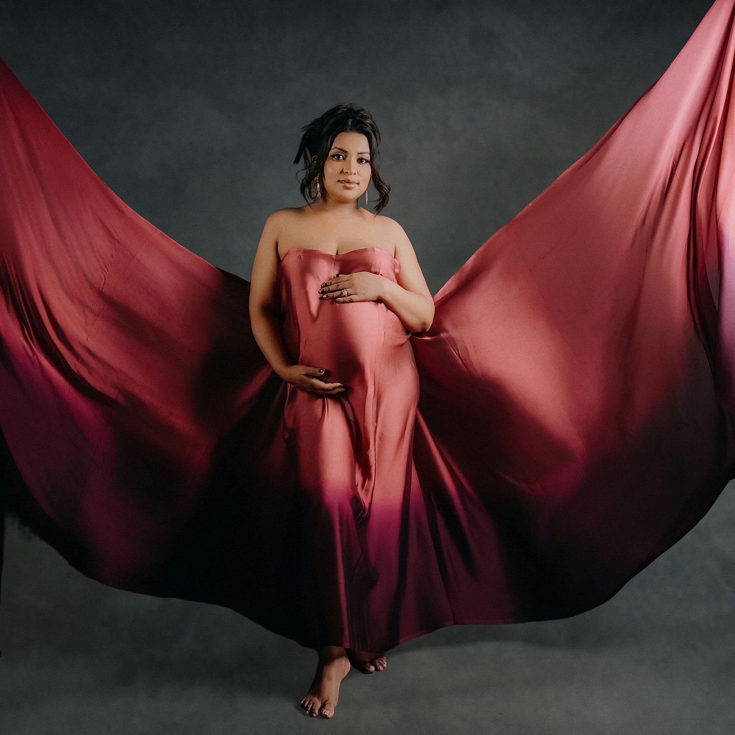 Pregnant woman in flowing red ombre gown with dramatic fabric wings spread wide, posing gracefully in a studio maternity portrait.
