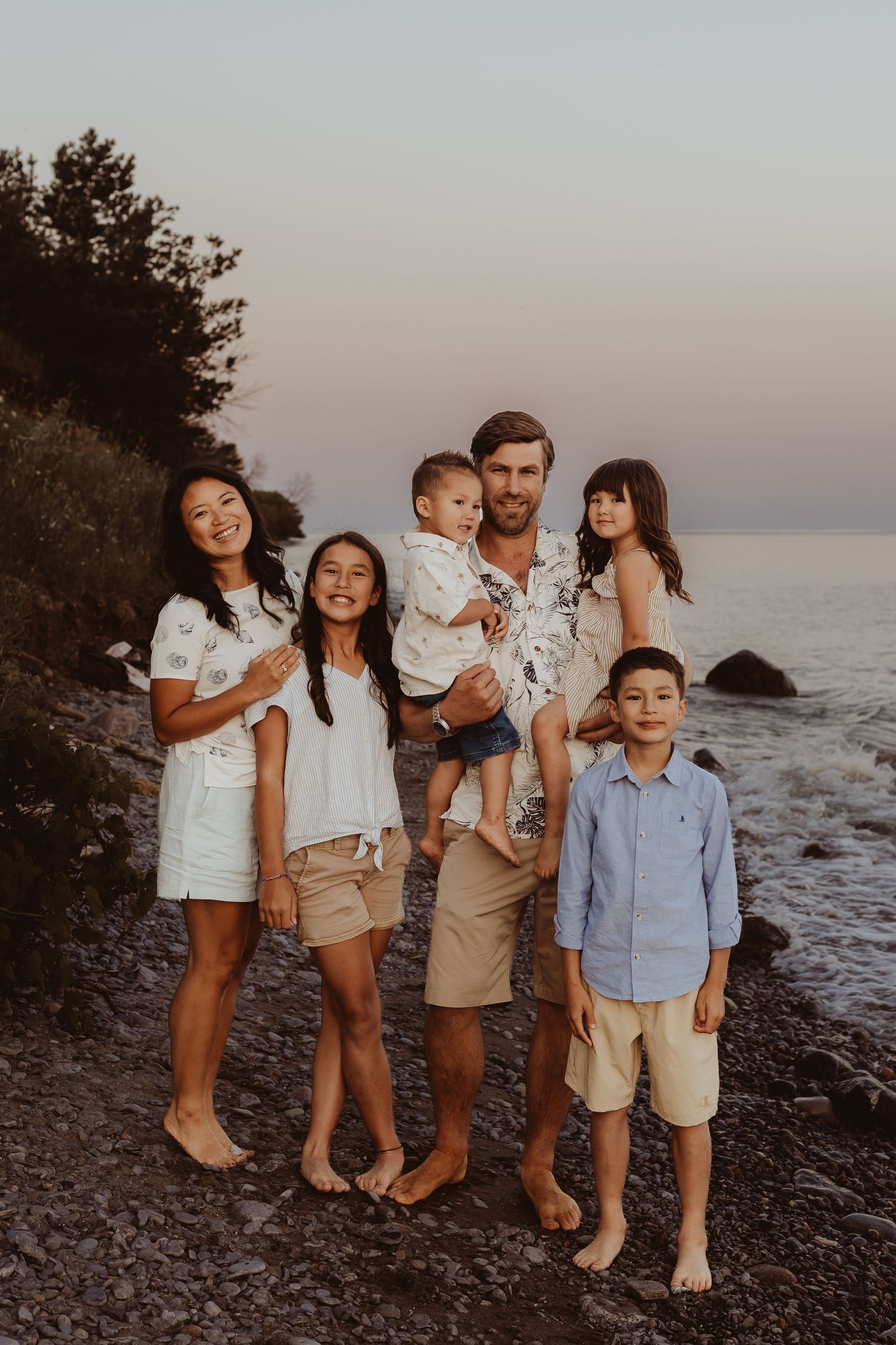 Happy family of six posing outdoors at golden hour, parents with four children smiling in a lush green park setting.