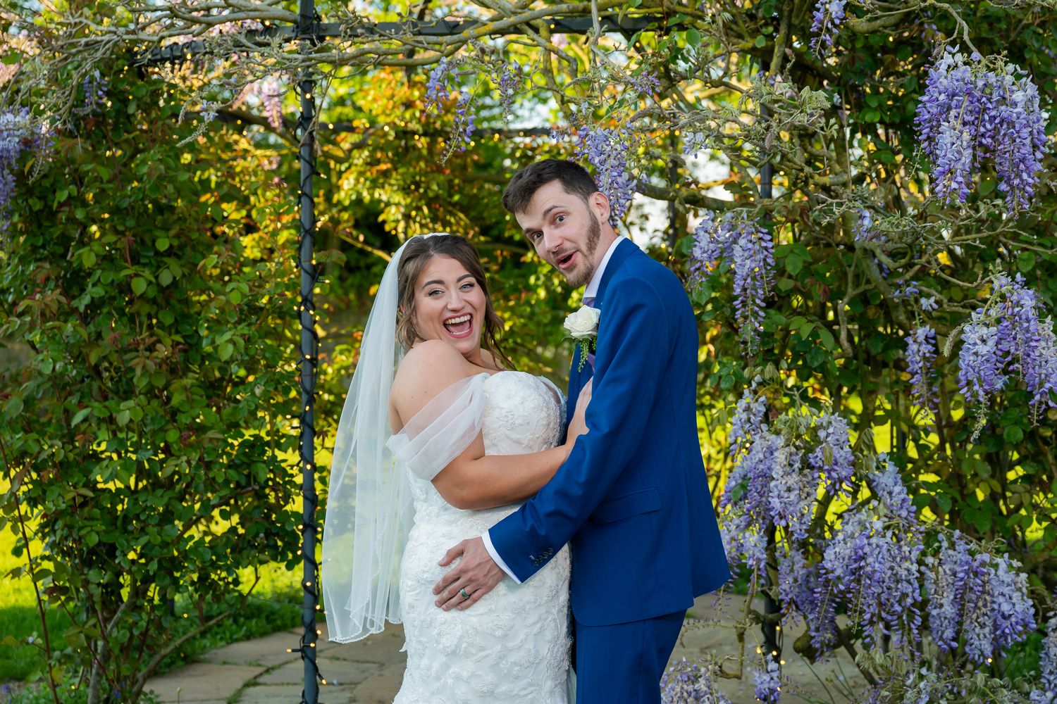Bride and groom smiling under a wisteria-covered garden arch, bride in white lace gown with veil, groom in blue suit.