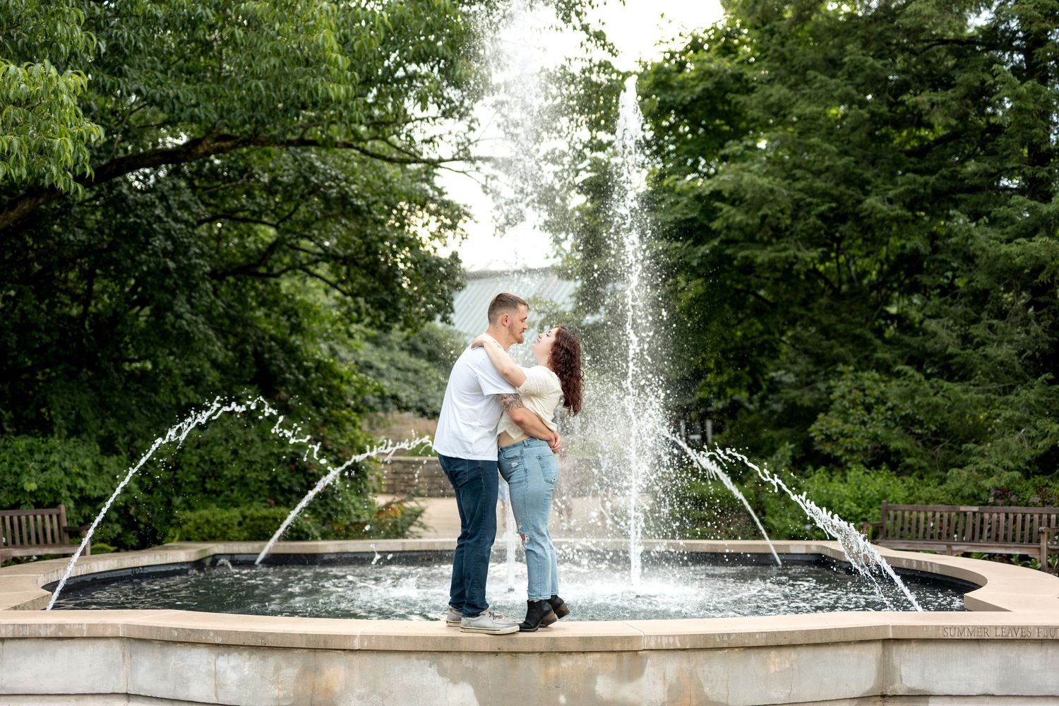 Couple embracing in front of a fountain in a lush green park, romantic outdoor engagement photo.