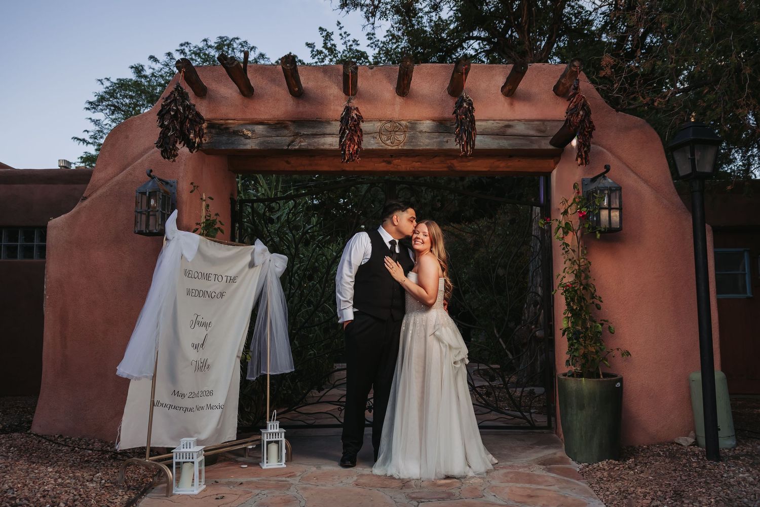 Couple sharing a romantic kiss under a rustic adobe pergola at dusk, with wedding dress displayed nearby.