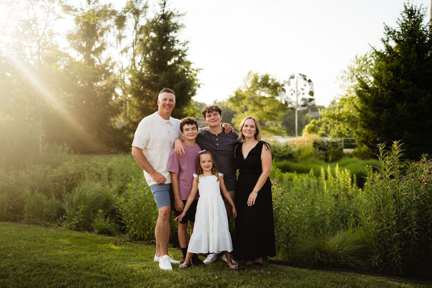 A joyful young girl in a white dress laughs surrounded by smiling family members outdoors in warm sunlight.
