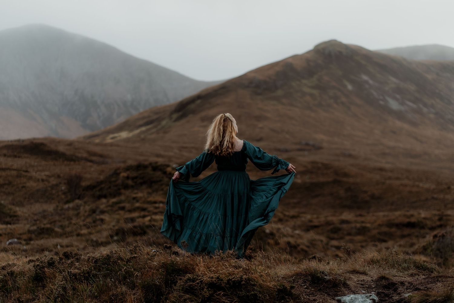 A figure in a flowing teal dress stands on a misty mountainside overlooking rugged terrain at dusk.