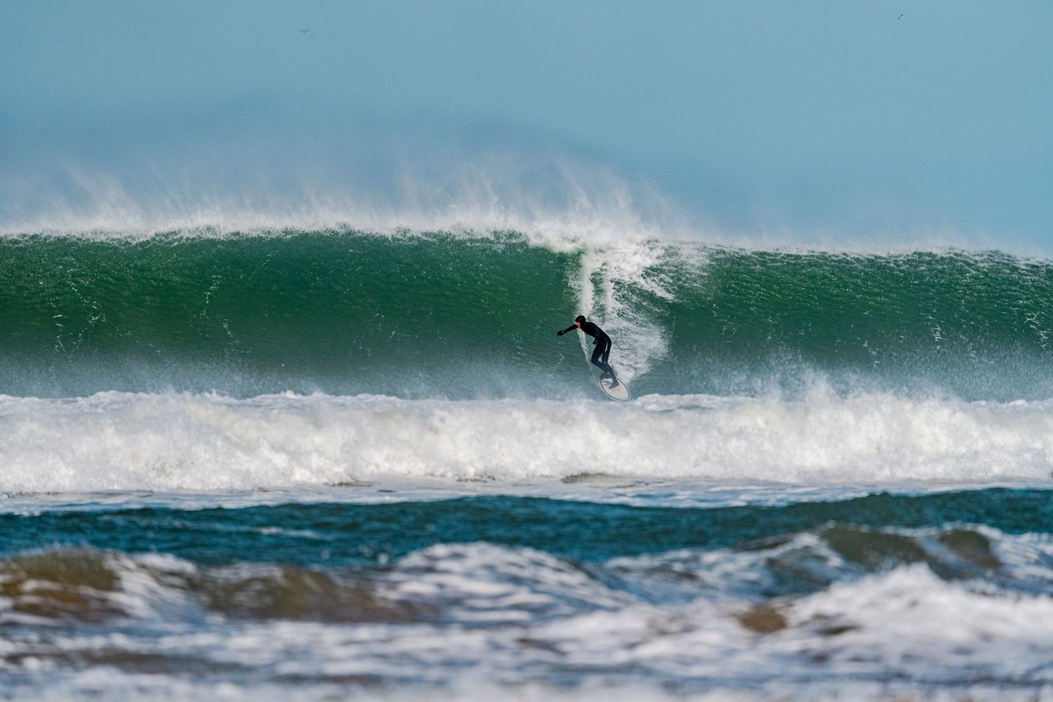 A surfer rides down a large green wave in rough ocean conditions during a dramatic surfing session at Fistral Beach