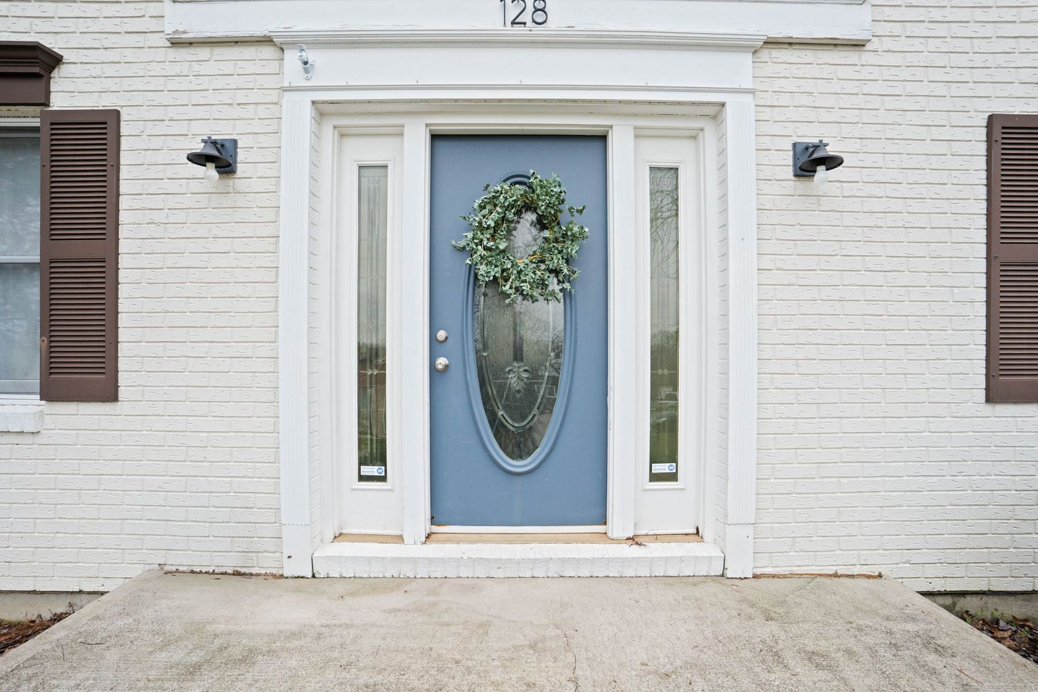 Blue front door with oval glass panel and wreath at house number 128 with white brick exterior.