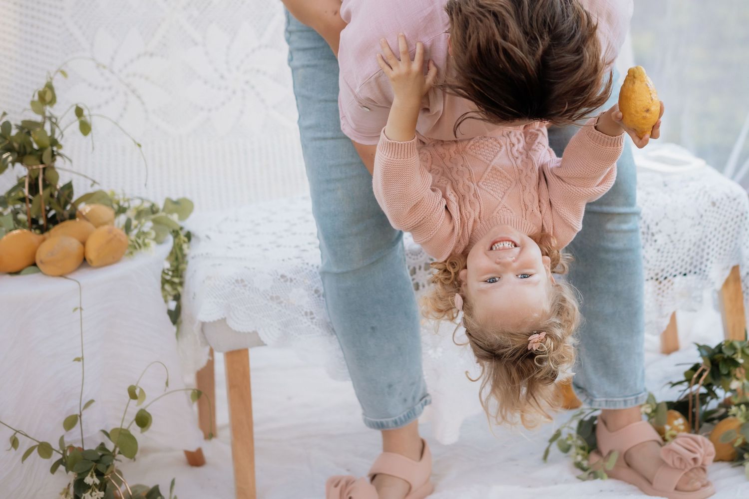 A person in jeans holds a child upside down while playing together on a white floor with citrus fruits nearby.