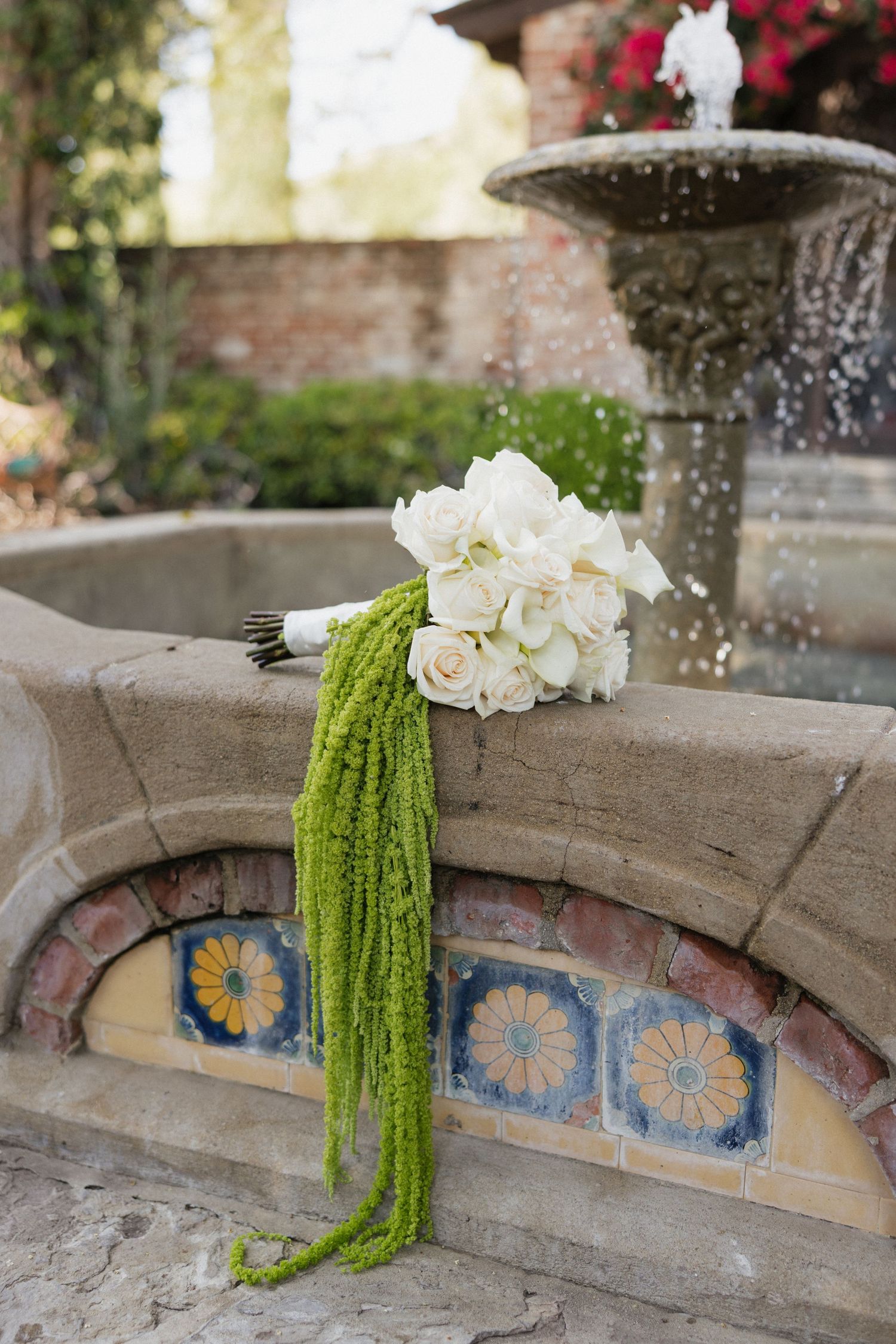 White rose and orchid bridal bouquet with green amaranthus on ornate tiled fountain in garden.