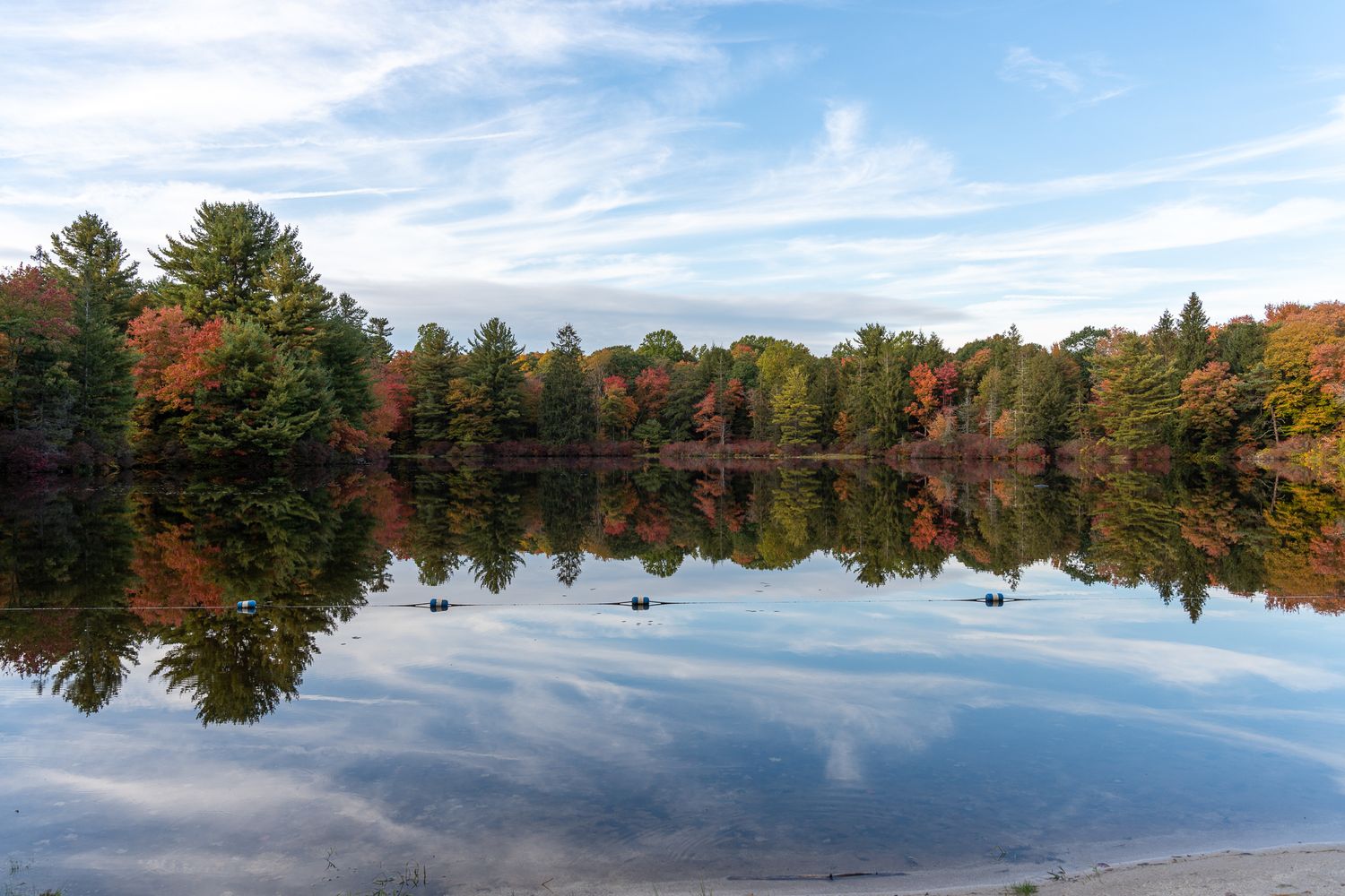 Calm lake reflecting colorful autumn trees with red, orange, and green foliage under a partly cloudy blue sky.