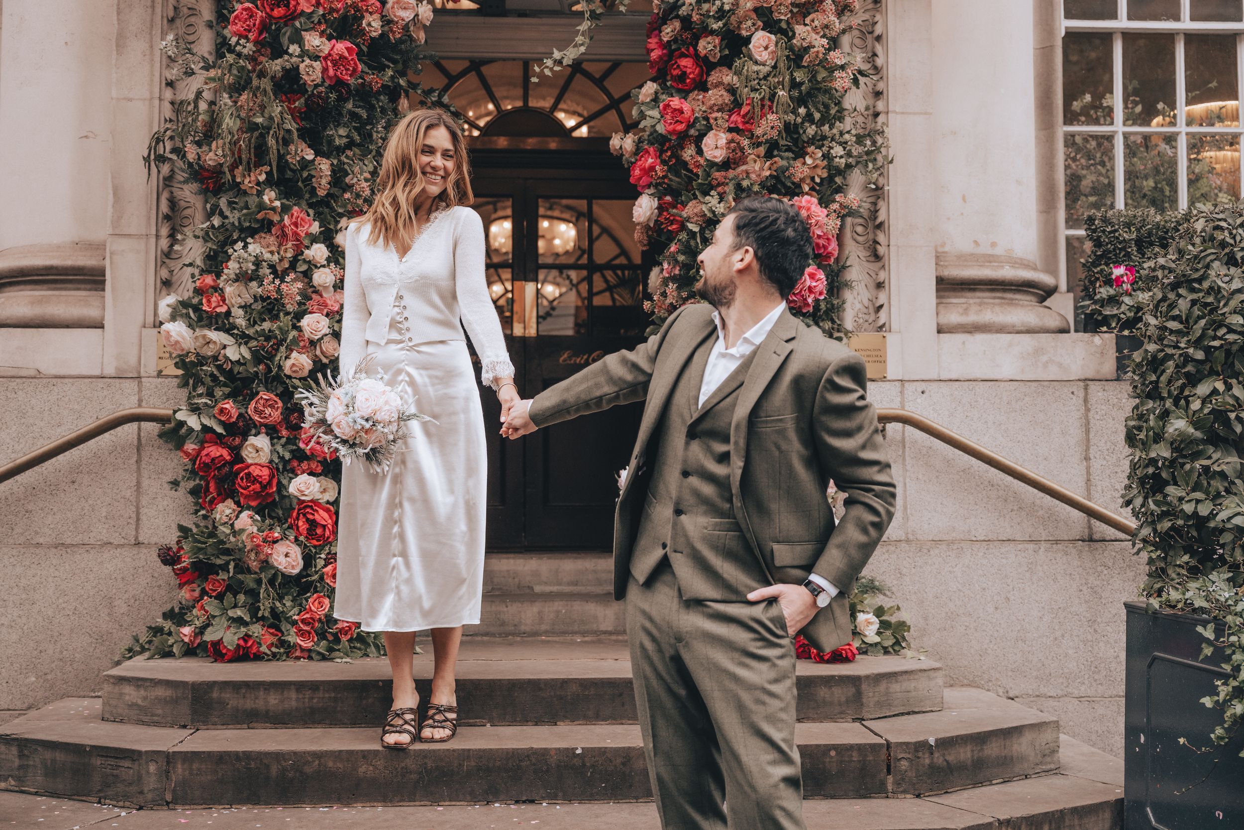 Bride and groom holding hands on steps adorned with lush floral arch of red and pink blooms at elegant venue.