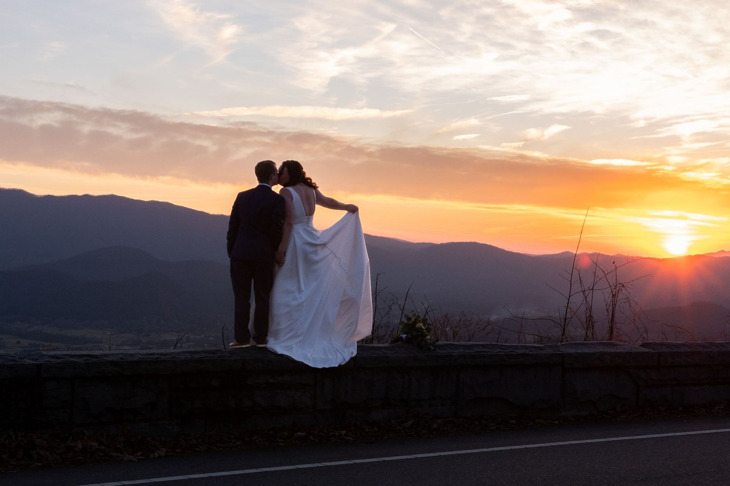 A silhouetted couple embraces on a mountain road against a dramatic sunset sky with mountain ranges in the background.