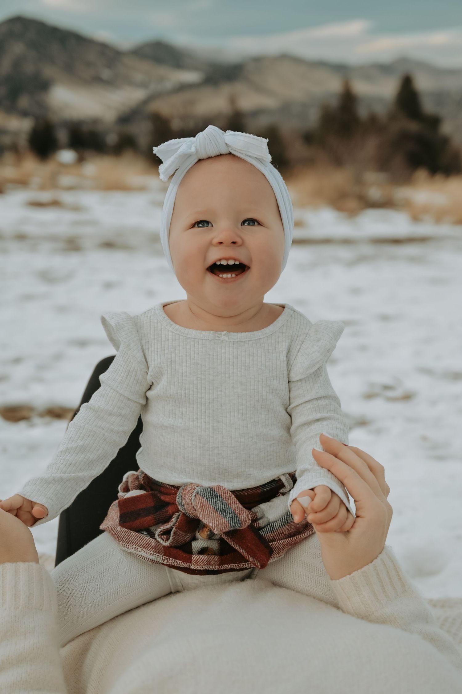 Laughing baby girl in white ruffle top and blue bow headband sitting outdoors in snowy winter landscape in chatequa park boulder, CO
