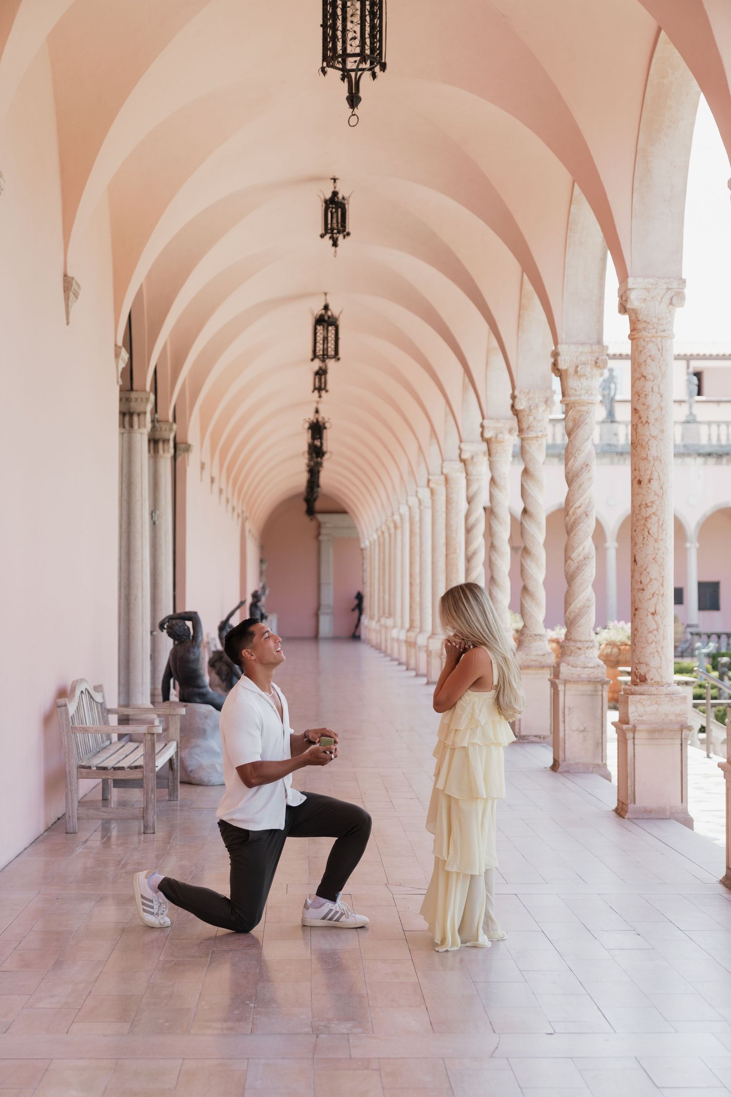 Man proposing on one knee to woman in elegant pink arched colonnade corridor.