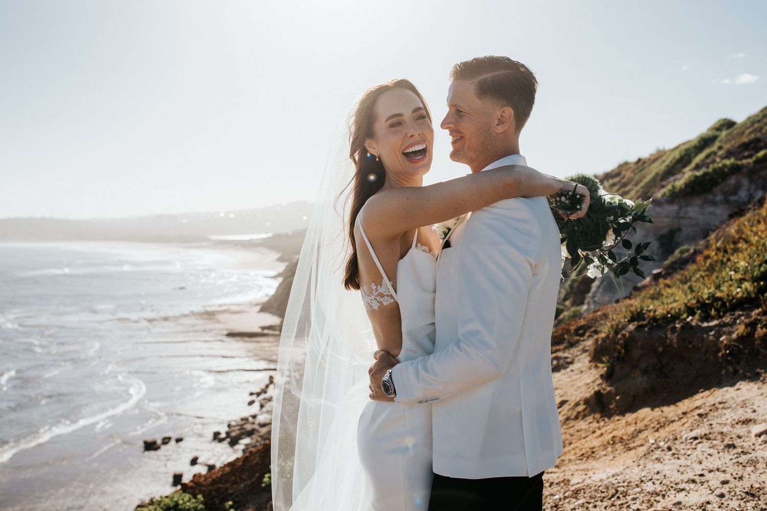 Happy couple in wedding attire embracing on a scenic coastal cliff with ocean views on their wedding day.