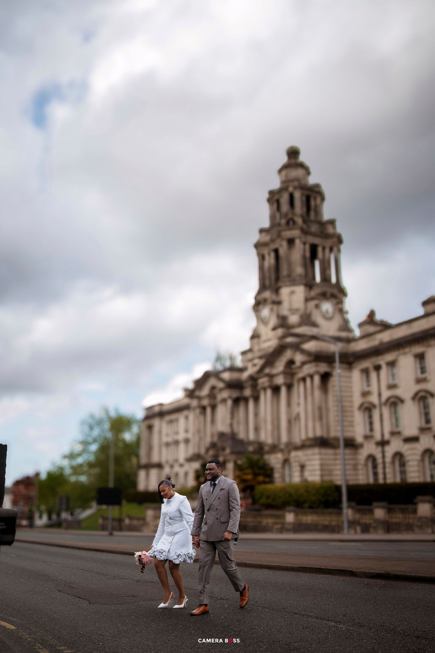 A wedding couple walks in front of a grand historic building under a cloudy sky.