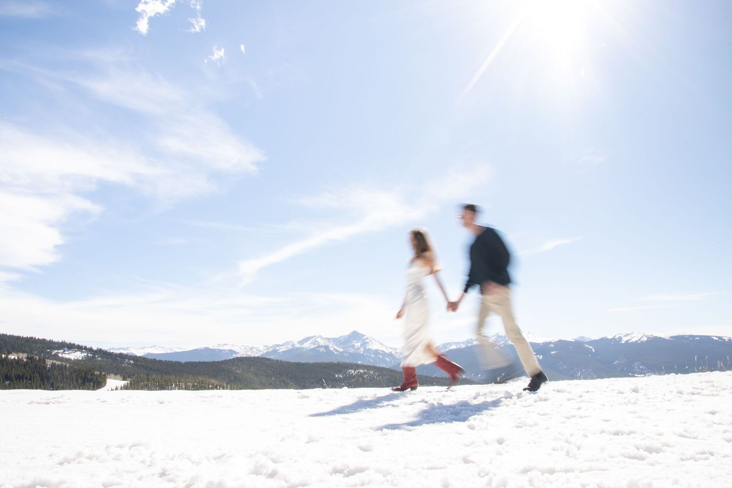 Couple holding hands running through snowy mountain landscape under bright blue sky in winter wedding attire in Vail Colorado engagement.