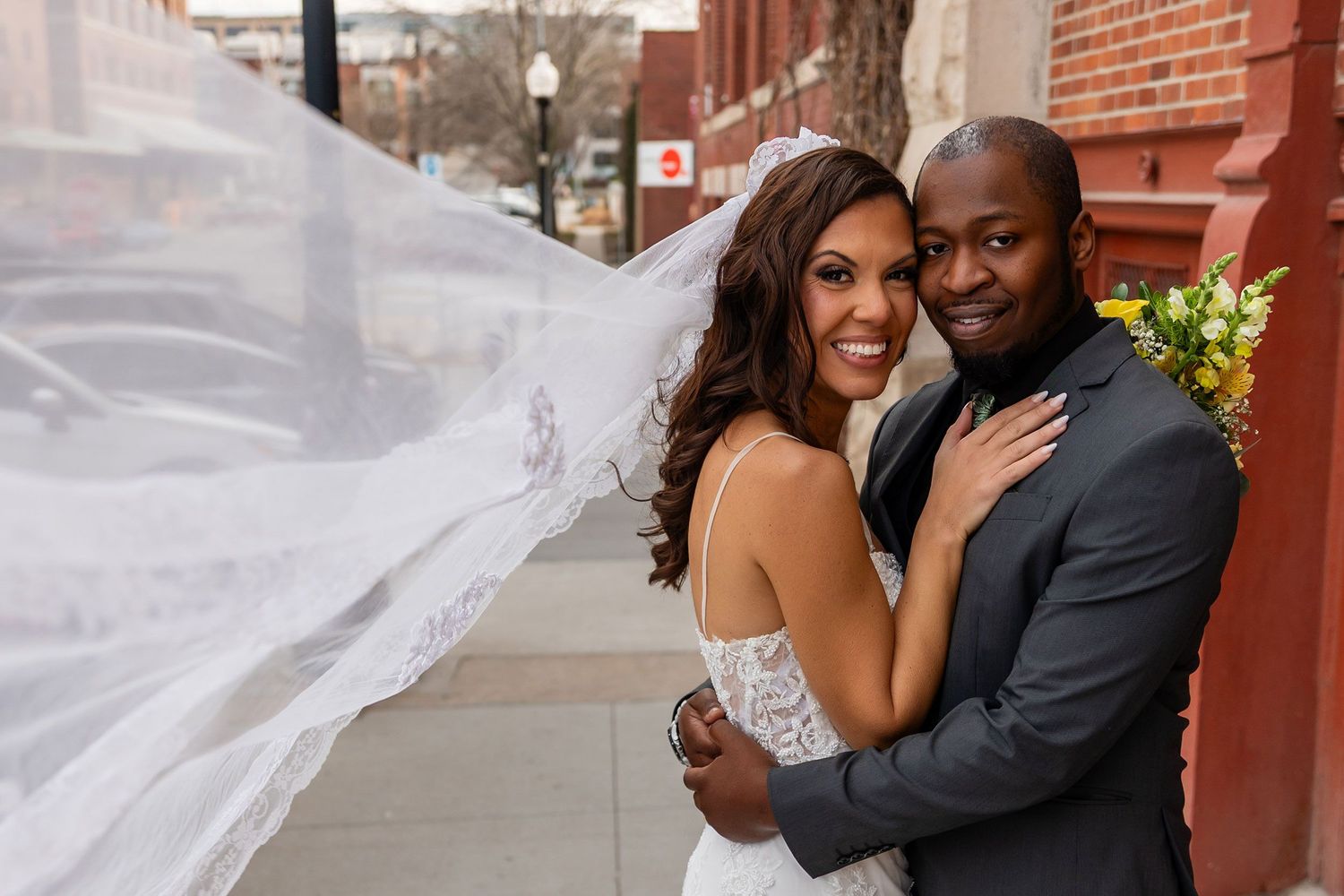 Bride and groom pose outdoors on wedding day, her veil flowing in the wind, holding a bouquet near a brick building.