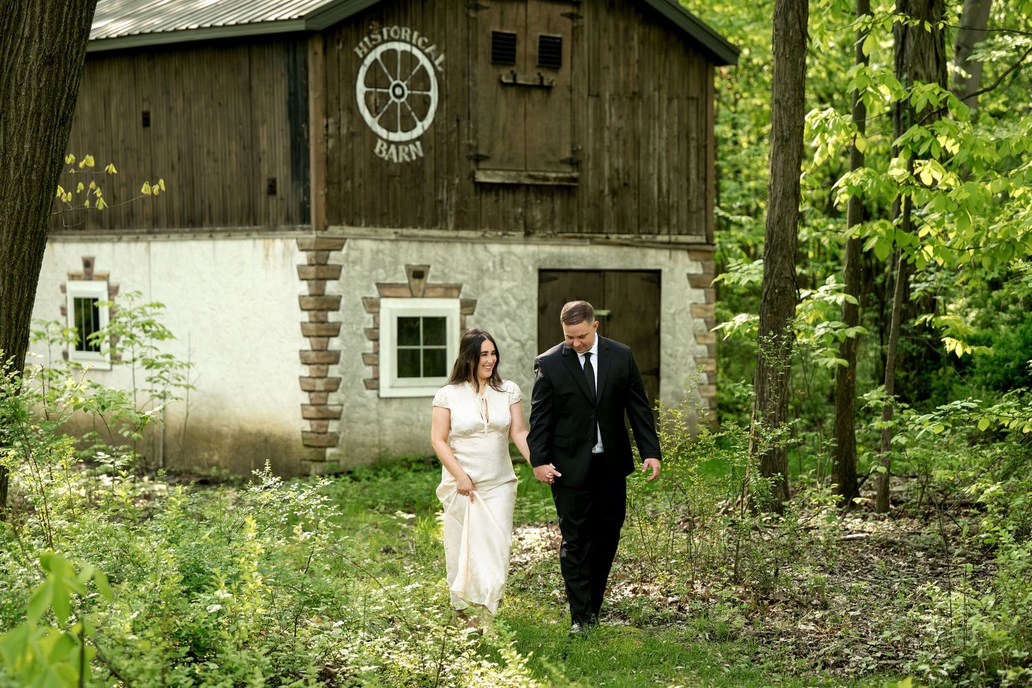 Couple in formal attire walking hand in hand near a rustic barn surrounded by lush green trees.