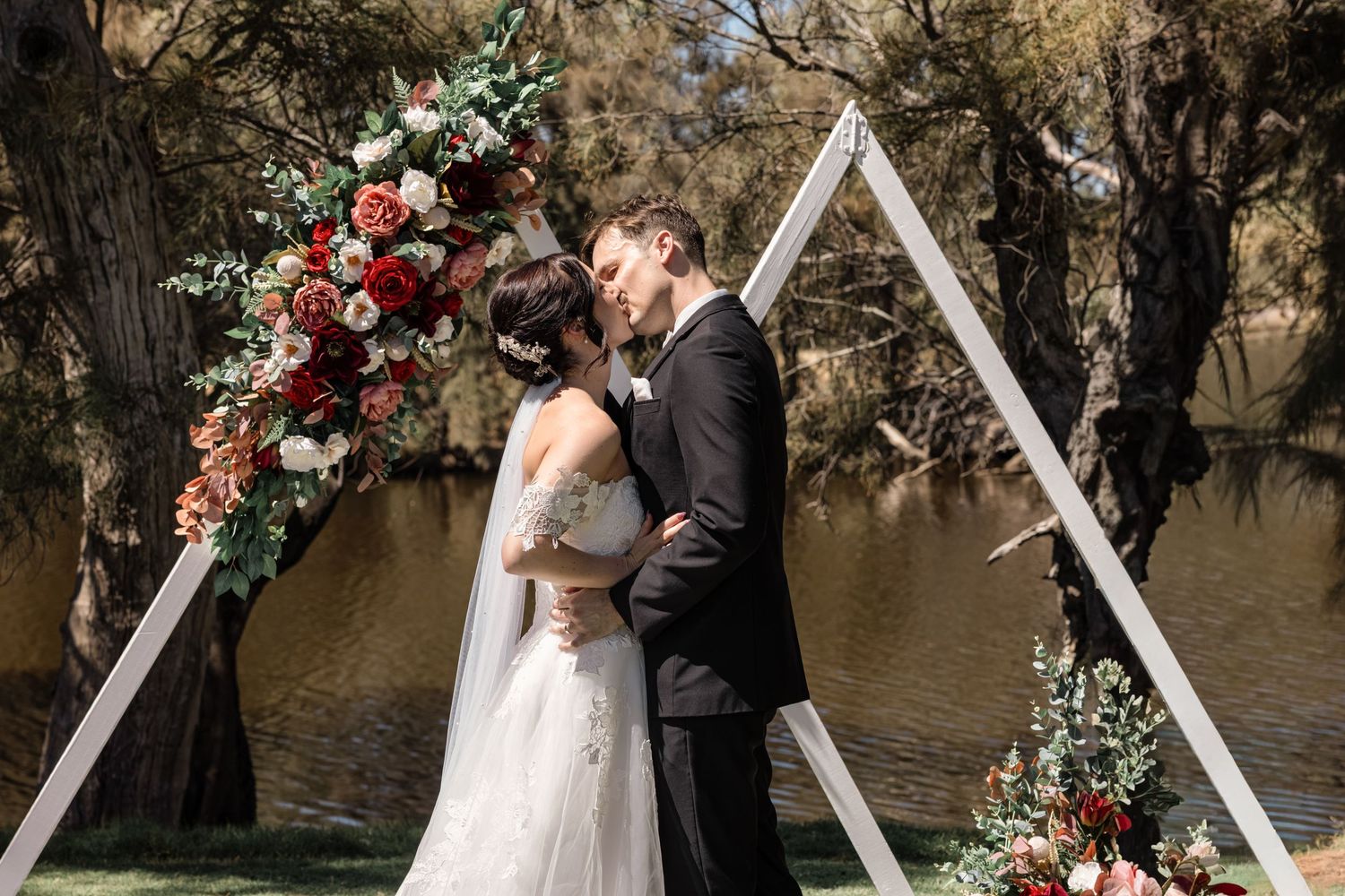 Bride in white gown and veil adjusts groom's tie outdoors at rustic countryside wedding venue.
