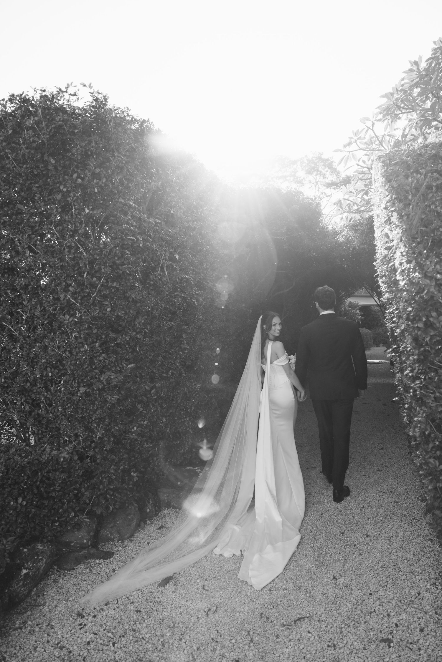Bride in white off-shoulder gown with long veil and groom in black tuxedo pose outside elegant venue.