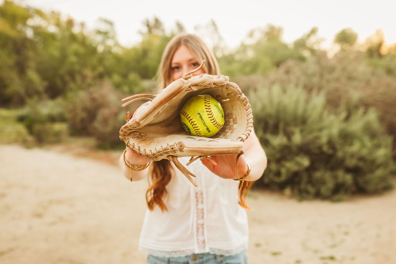 Young girl holding a softball glove with a yellow softball outdoors in a natural setting.