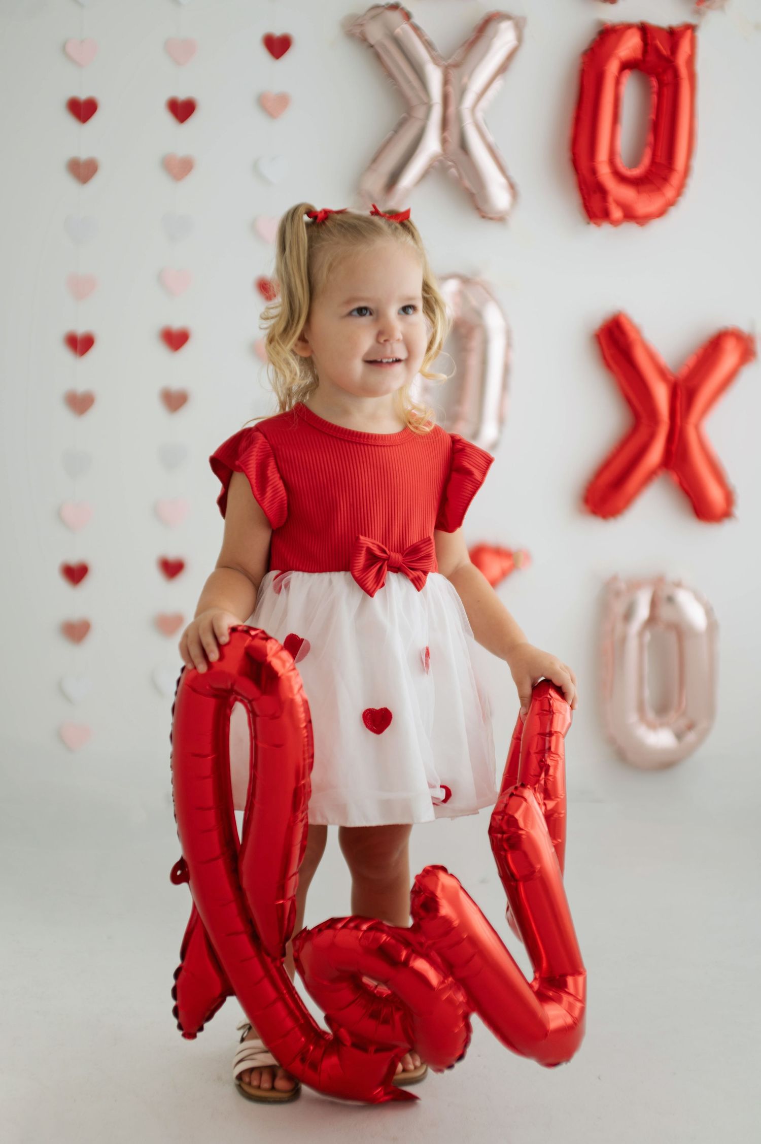 Toddler girl in red and white dress holds LOVE balloon for Valentine's Day photo with XOXO balloons.