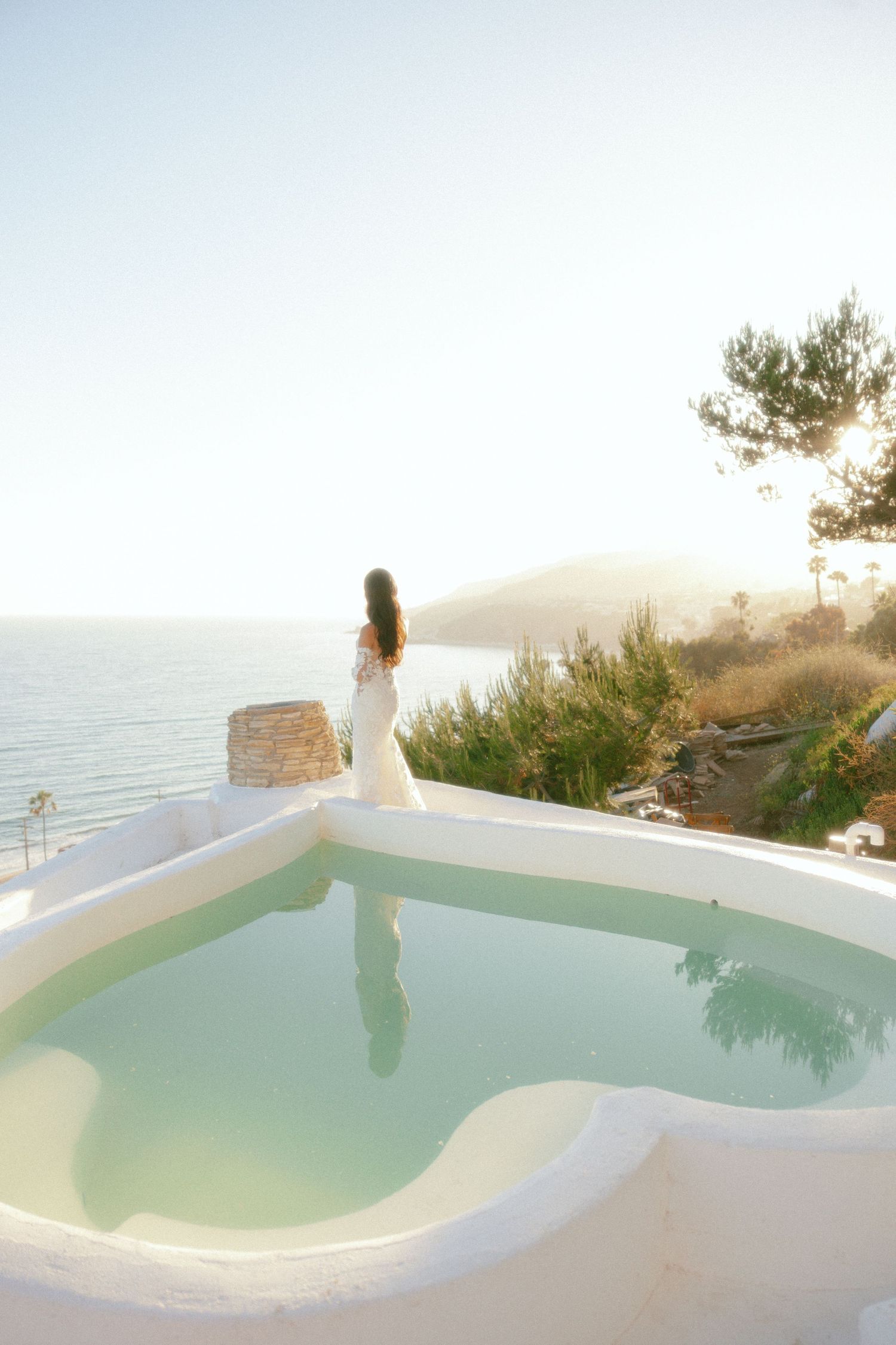 Woman in white dress stands by a turquoise pool with stunning Mediterranean sea views on a sunny day.