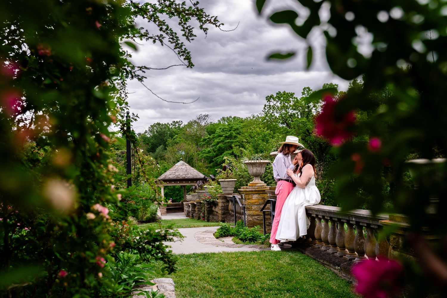 Couple sharing a romantic kiss in a lush garden with stone architecture and blooming pink flowers on a cloudy day.