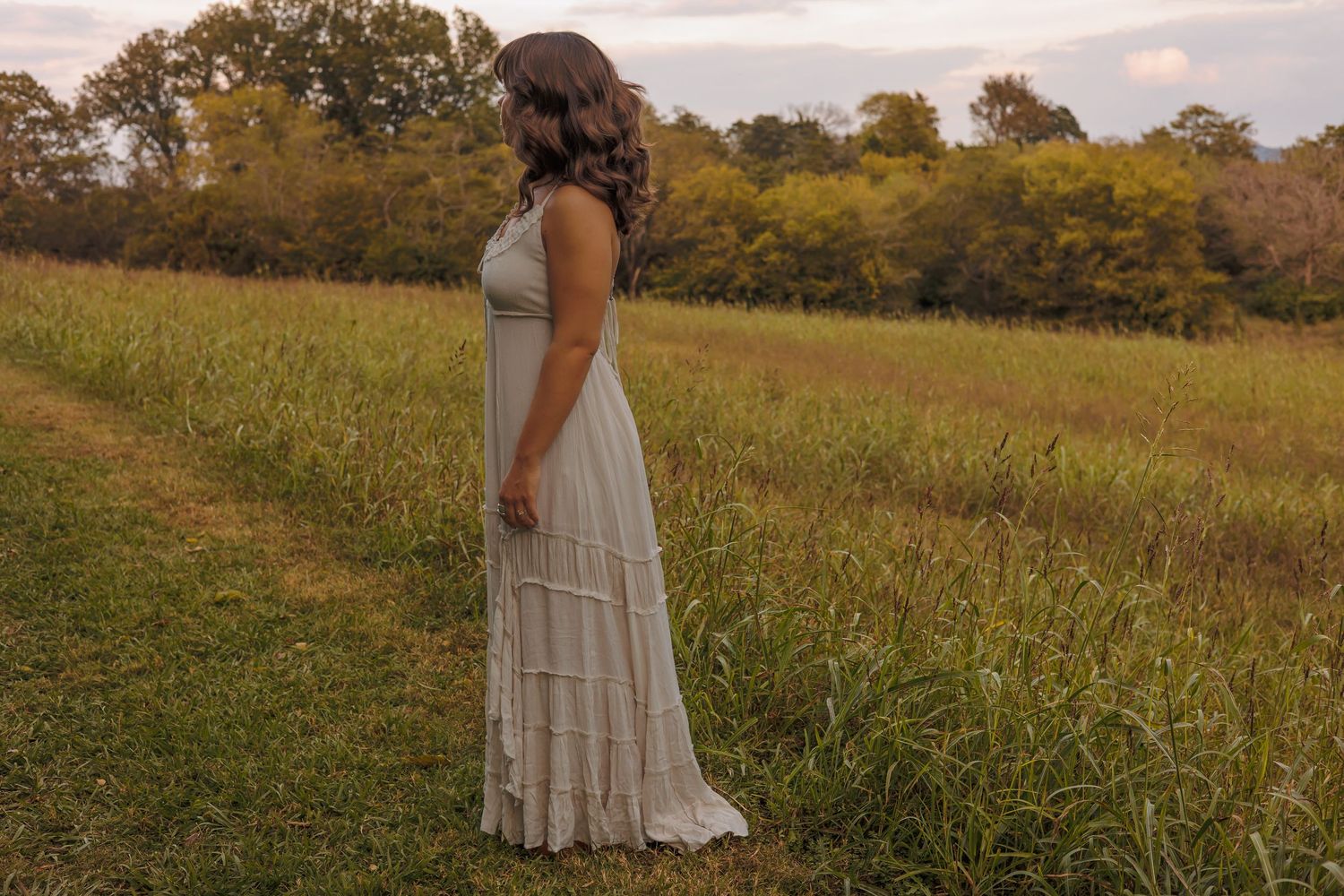 A person in a flowing white dress stands in a golden meadow at sunset with trees in the background.