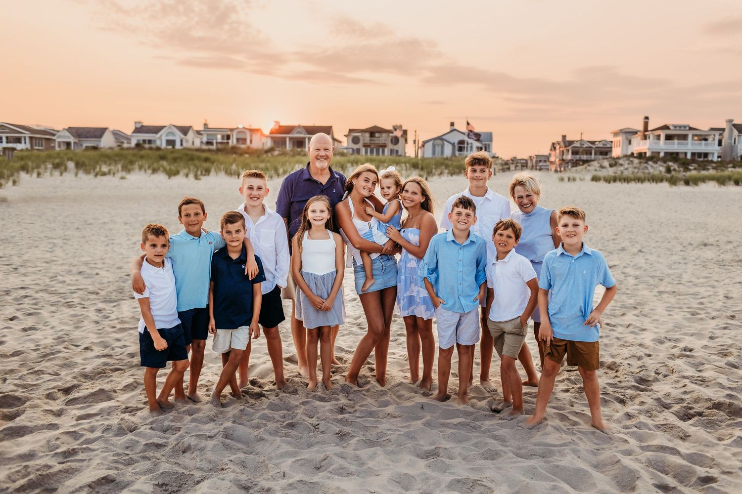 Large family group photo on a sandy beach at sunset with children and adults in coordinating blue and white outfits.