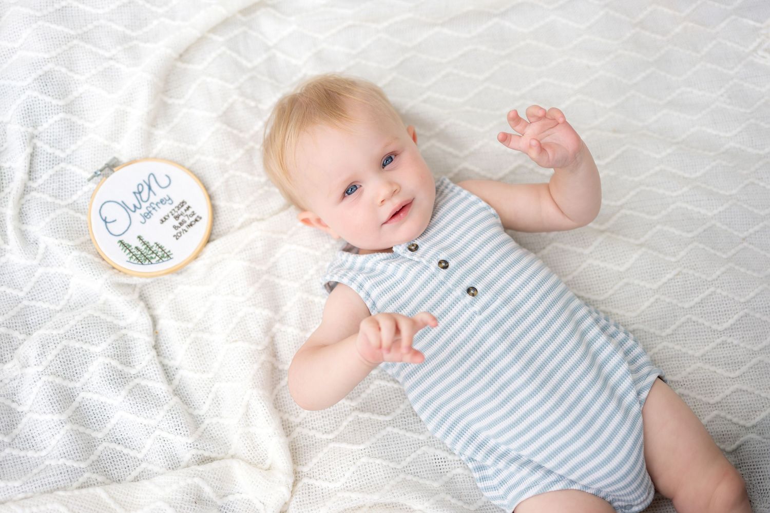 Smiling baby boy named Owen lying on white blanket wearing striped romper, reaching up with one hand.