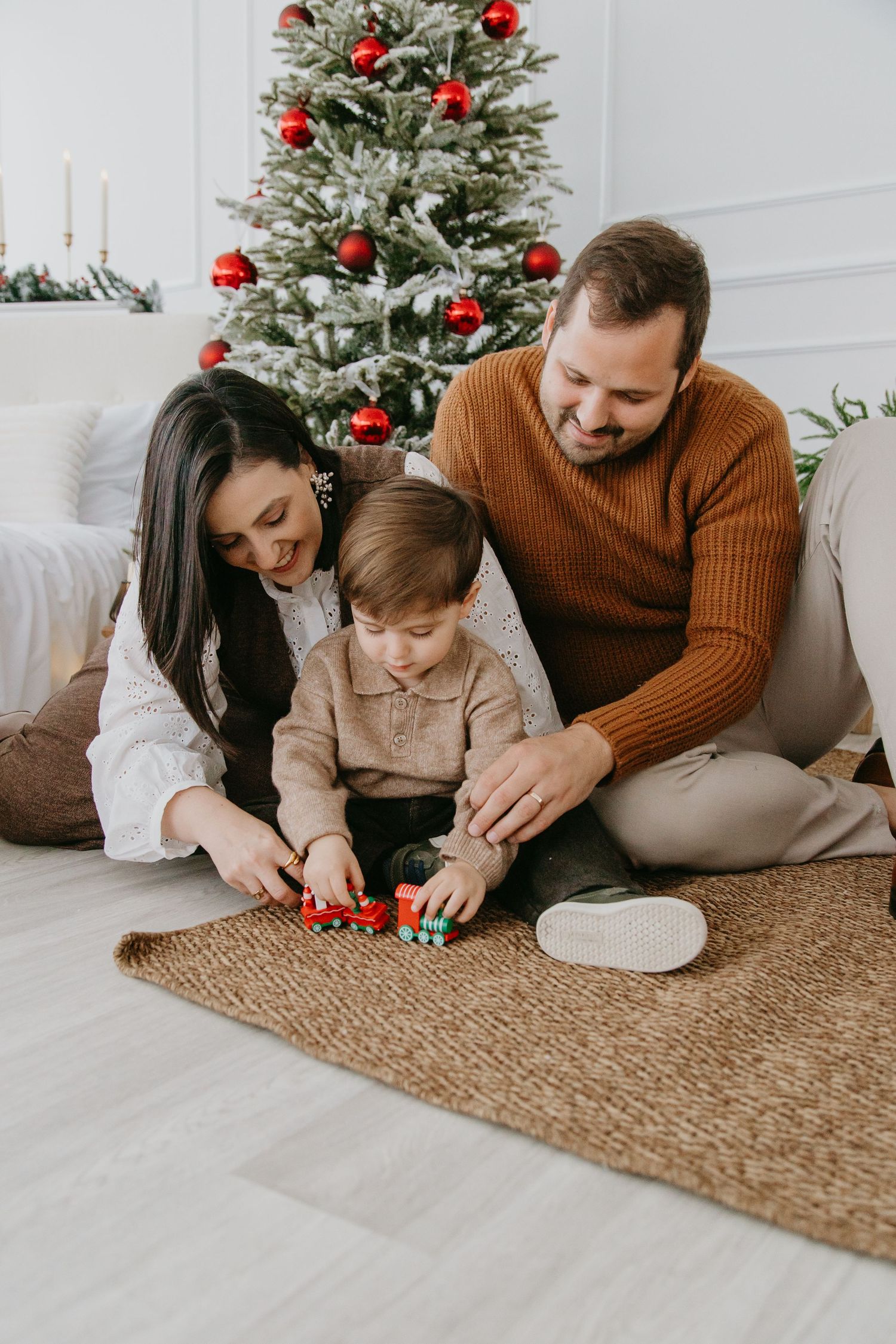 Family plays together on jute rug by decorated Christmas tree with red ornaments in bright modern living room.