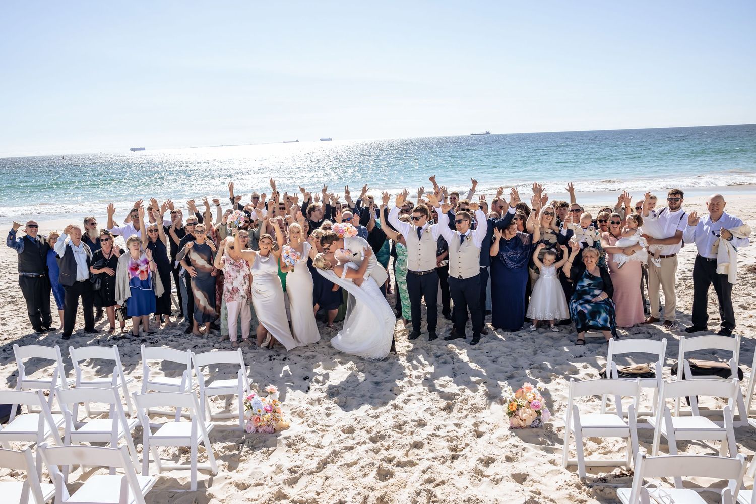 A large group celebrates on a sunny beach with white chairs set up for a wedding ceremony by the ocean.
