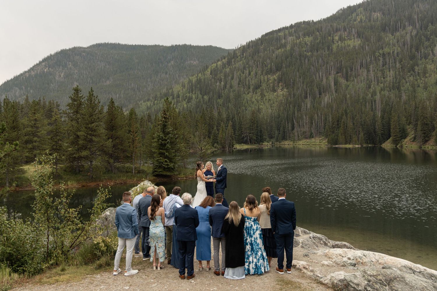 A group gathers by a tranquil mountain lake surrounded by evergreen forests.