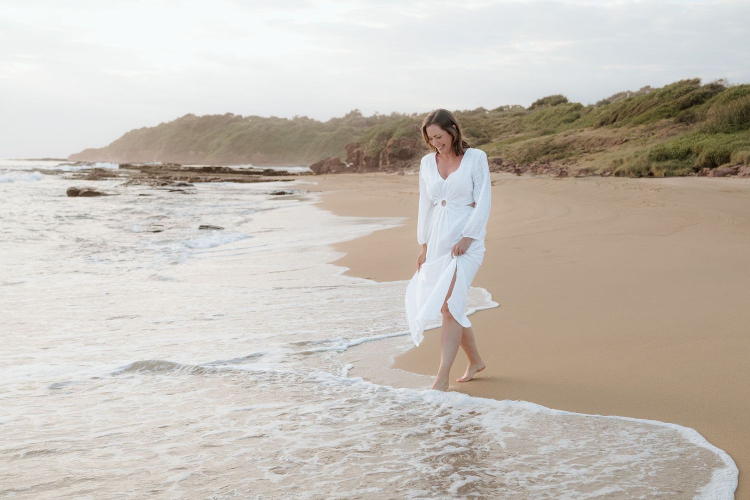 A person in a flowing white dress walks barefoot along a serene beach shoreline at sunset.