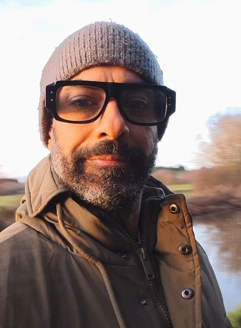 A person wearing a yellow knit hat and glasses smiles while taking selfies near a lake on a sunny day.