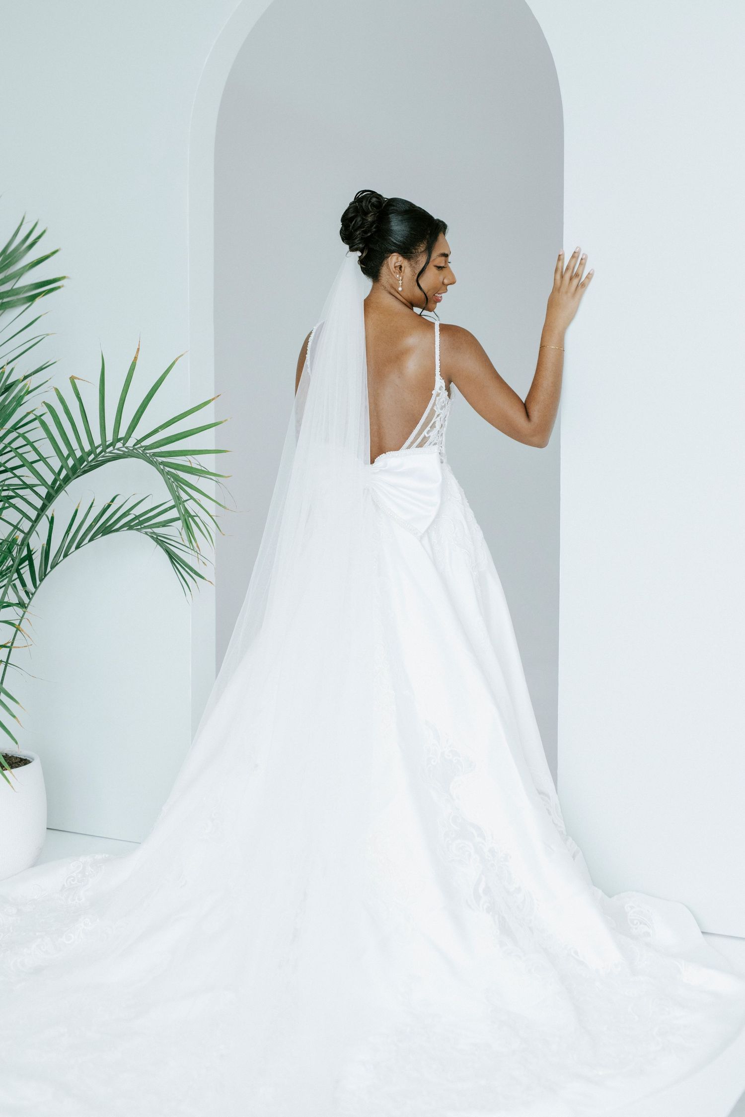 A bride in an elegant white wedding gown poses gracefully against a minimalist white backdrop with tropical palm leaves.