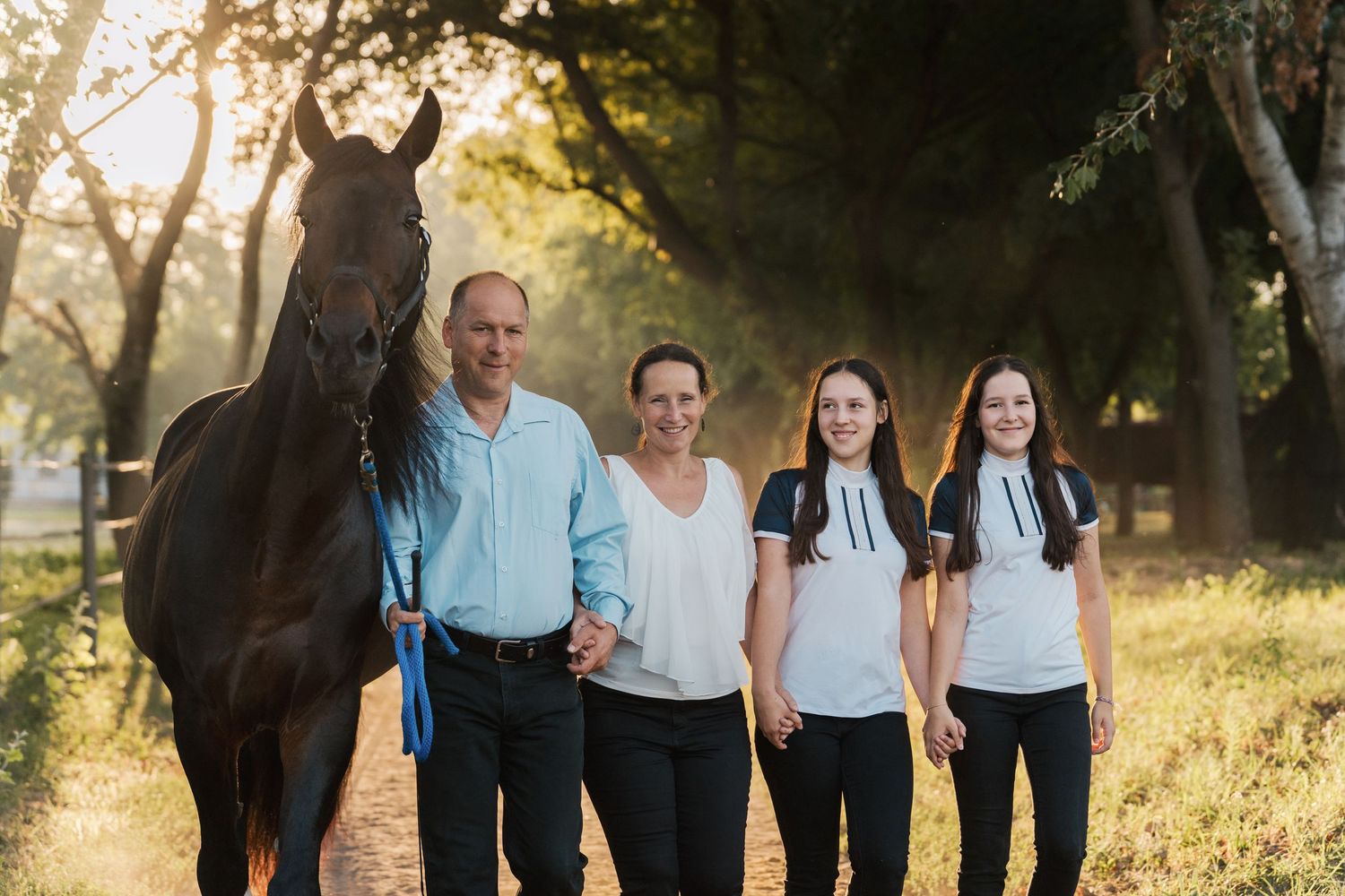 A family walks with their dark horse on a sunlit path through trees during a golden hour photoshoot in the morning in Sydney