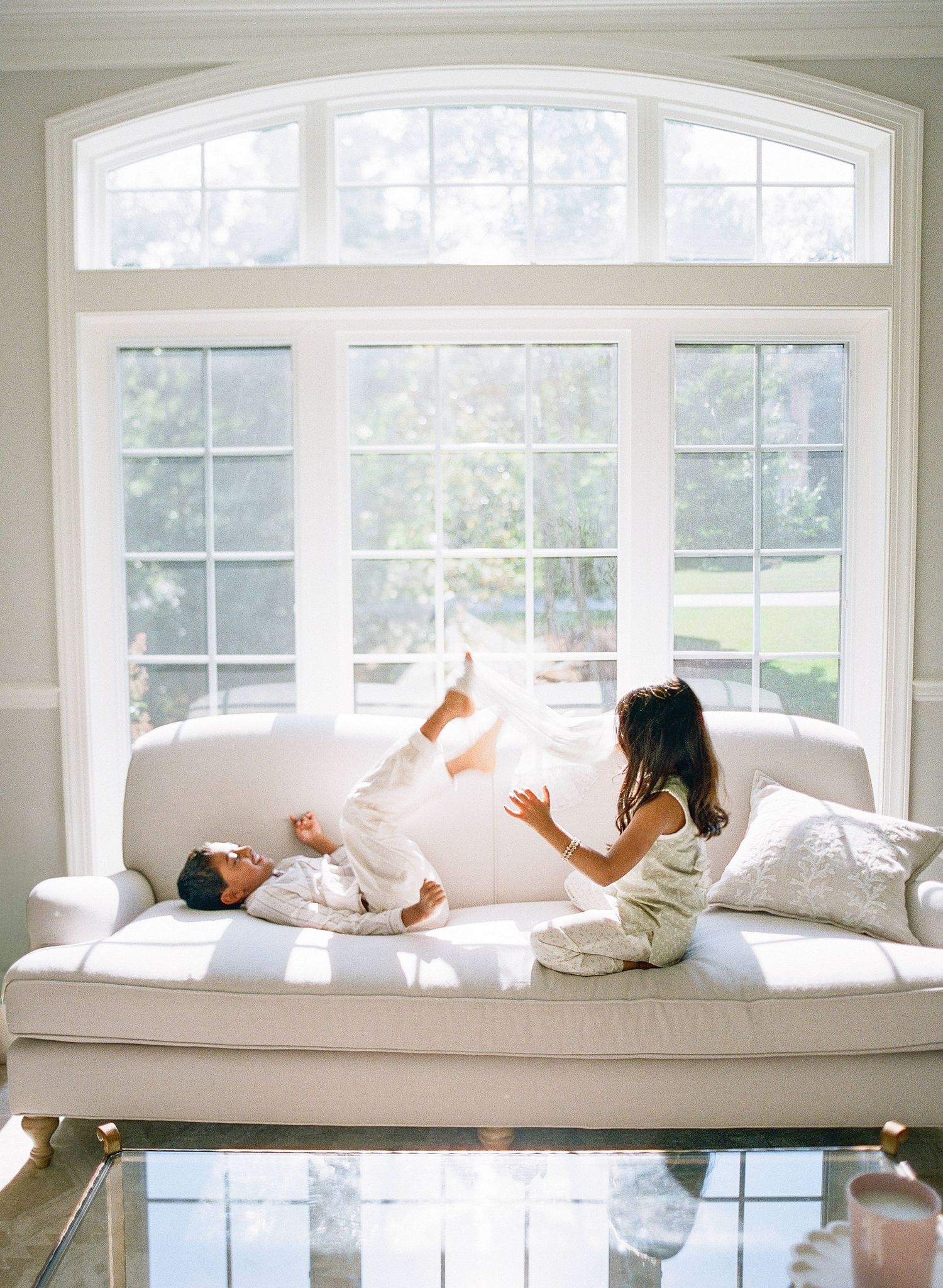 Two adults changing diapers of babies on a gray striped couch in a sunlit room.