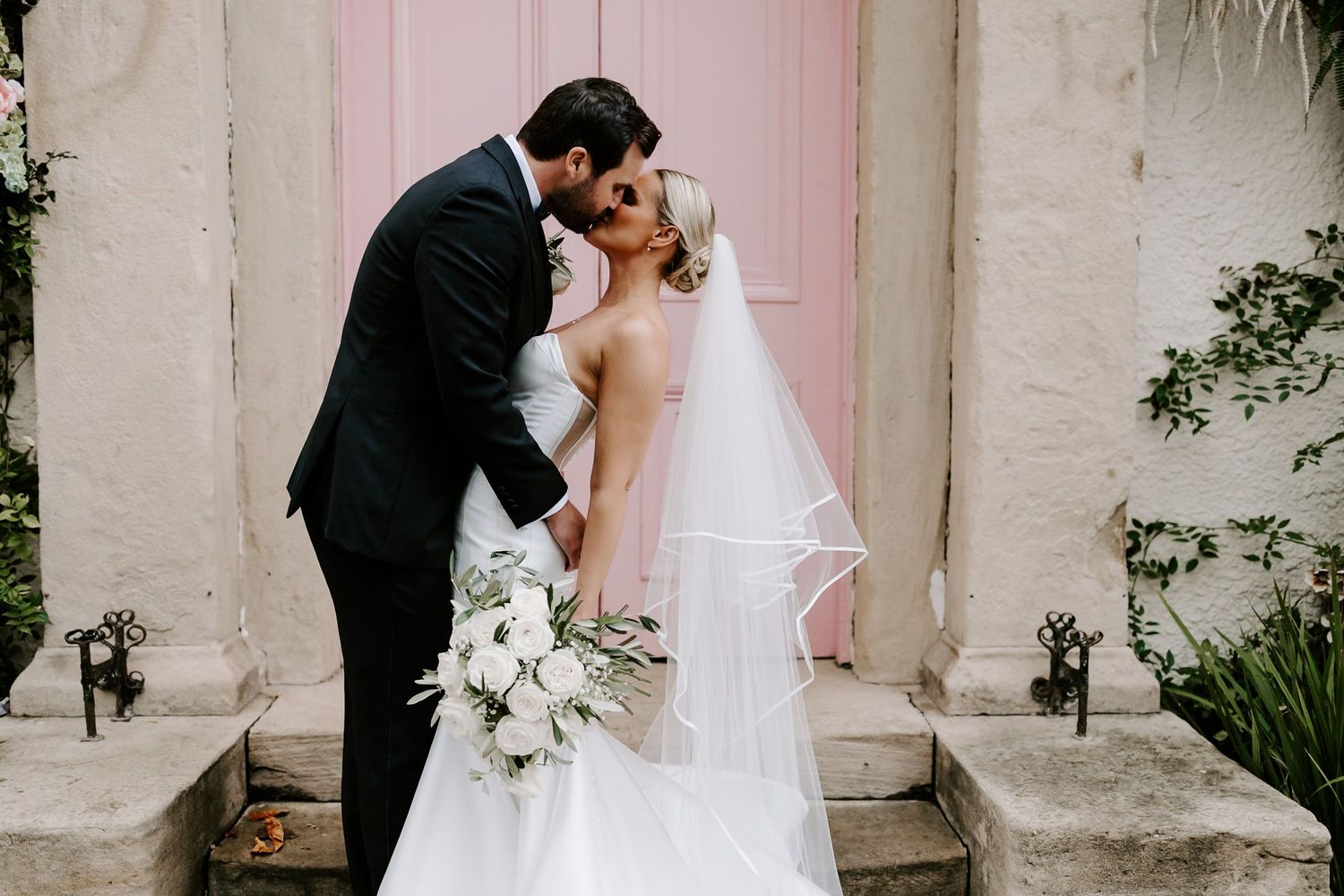 Bride and groom share a romantic kiss while holding a white floral bouquet against a white wall backdrop.