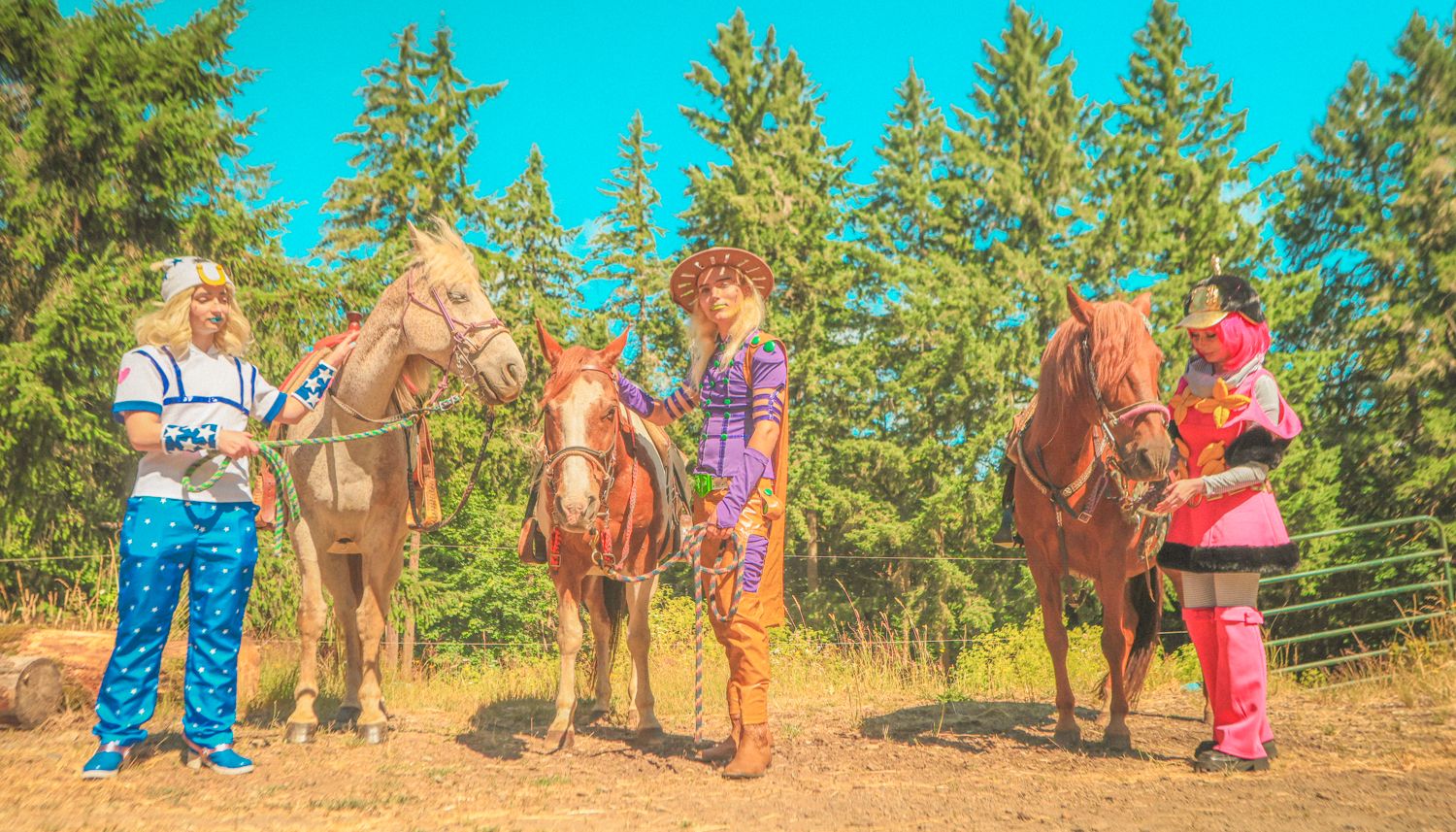 People stand alongside horses on a dusty trail with evergreen trees in the background on a sunny day.