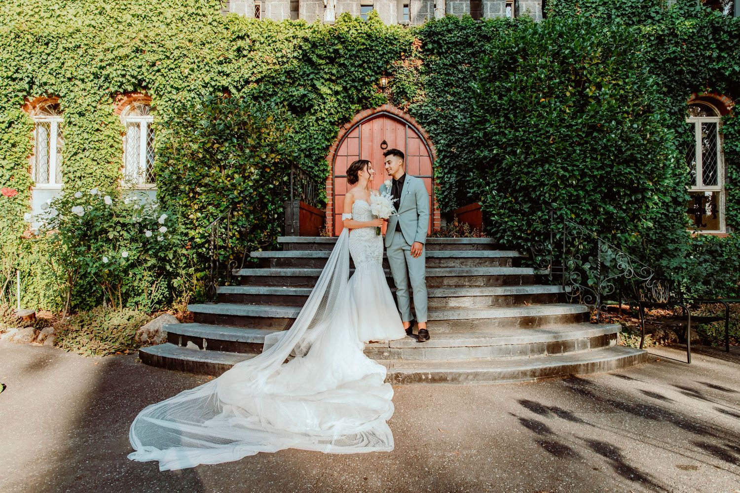 Elegant bride and groom pose by stone staircase surrounded by lush greenery and building in background.