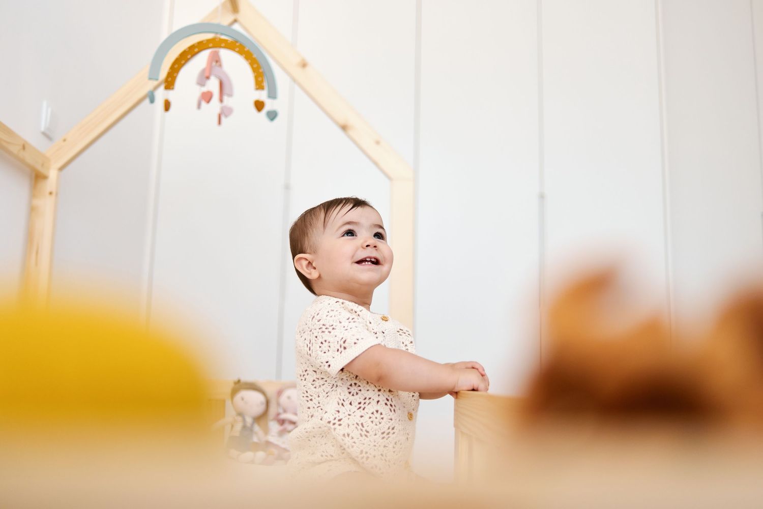 Adorable baby in a floral onesie standing in a wooden play gym looking up with a joyful expression.