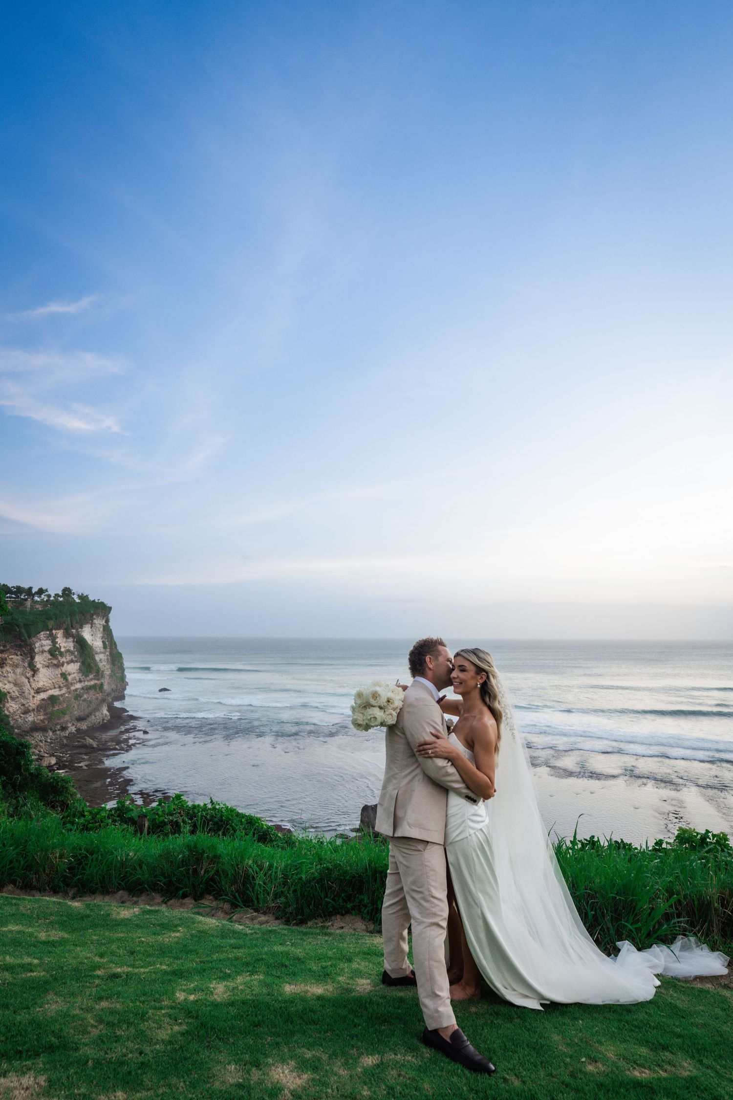 Silhouetted couple sharing a romantic kiss under flowing wedding veil at sunset with dramatic sky in background.