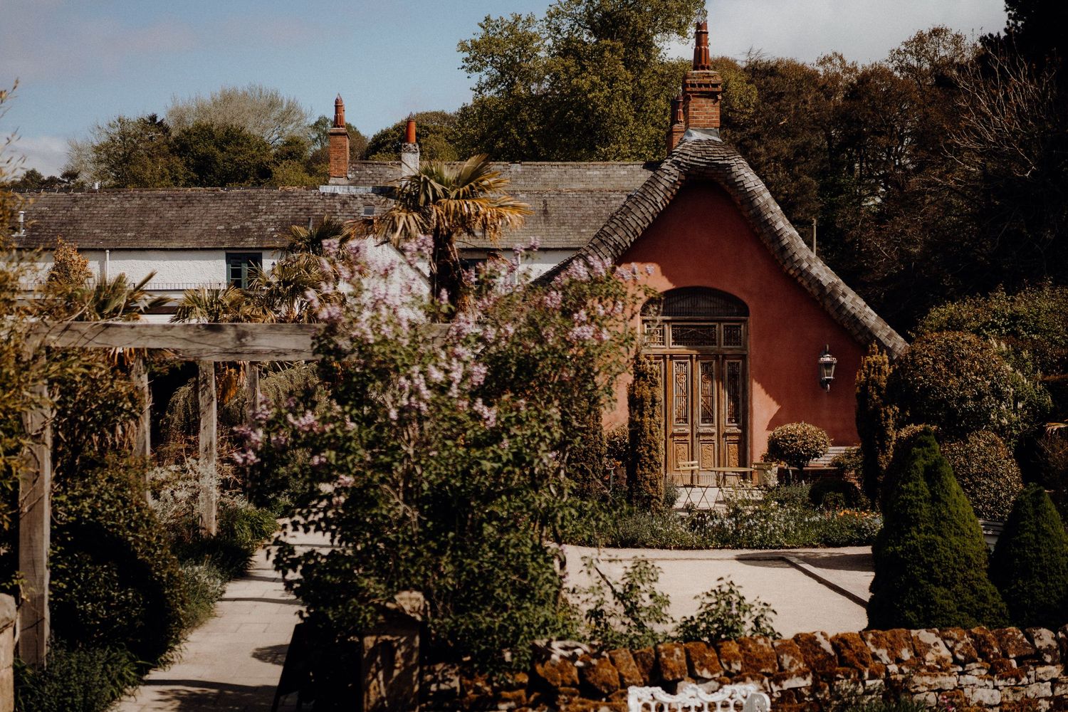 A charming red cottage with climbing roses and garden arbor in a rustic countryside setting.