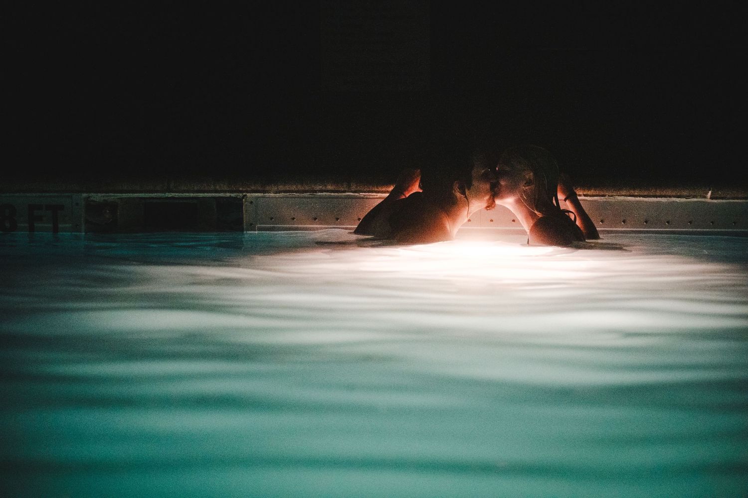 A glowing warm fire pit illuminates the surface of a turquoise swimming pool at night.