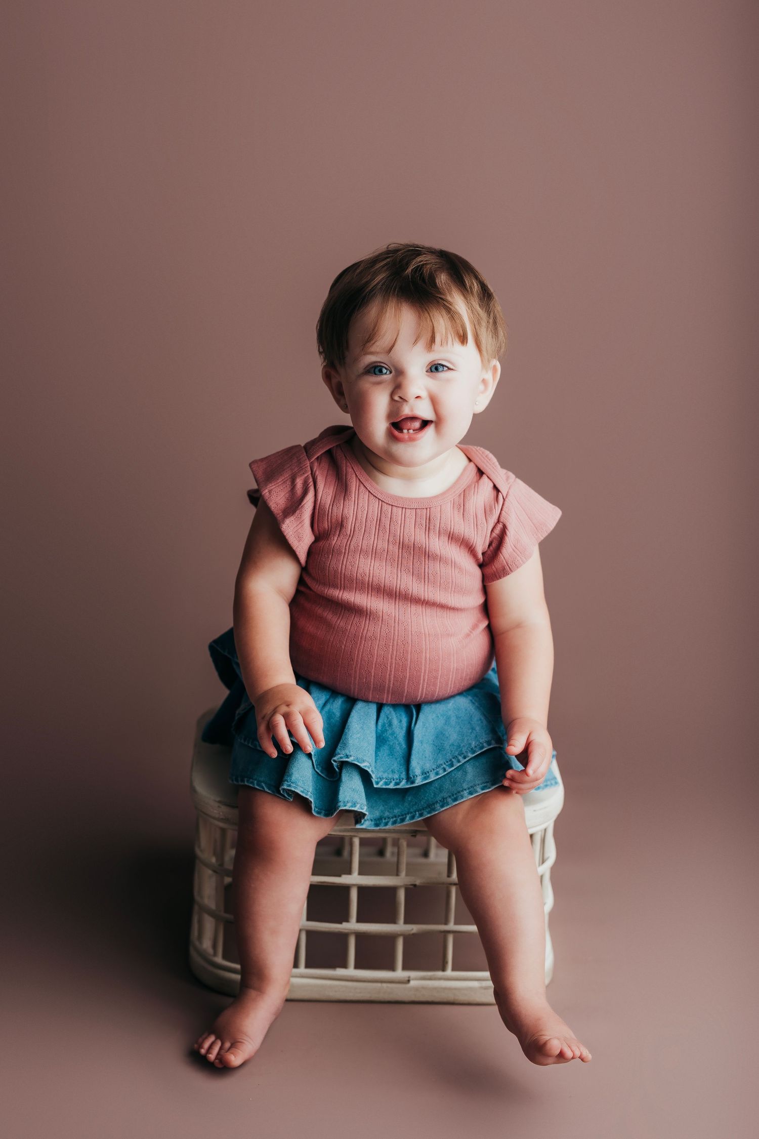 Child wearing pink ribbed top and blue tulle skirt sits on decorative block against dusky rose background.