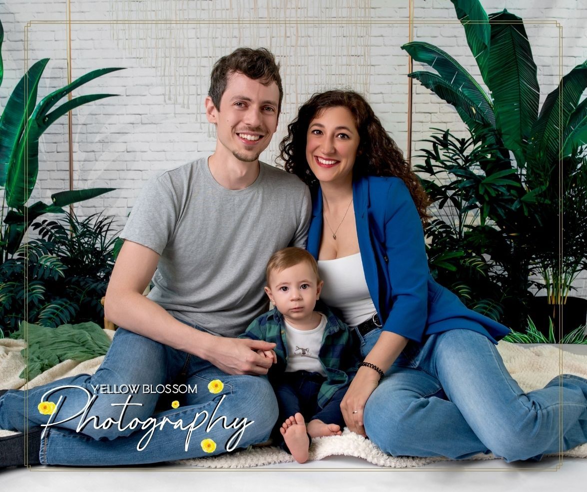 A family sits together against a brick wall with tropical plants, wearing casual clothing in blue, white and grey tones. Haverhill, Suffolk
